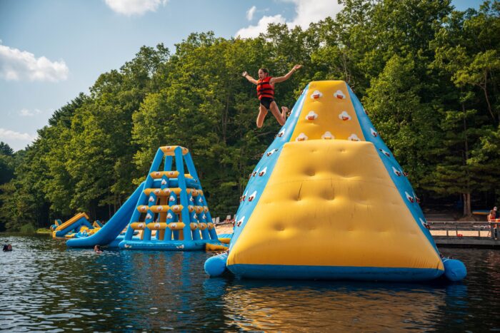 Person in red life vest jumping from yellow and blue inflatable climbing structure on a lake, surrounded by other water obstacles.